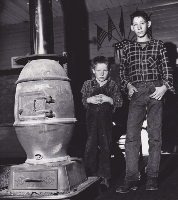 Two boys standing next to a furnace. The boys are dressed in overalls, jeans, and flannel. American flags can be seen in the background. 
